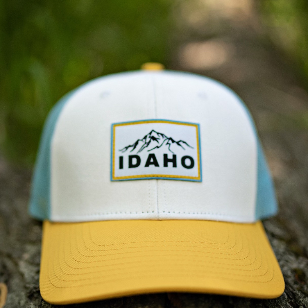 Modern Idaho Trucker Hat resting on a log, featuring a bold mountain patch and yellow brim—designed by TatorJo, a small Idaho apparel business.
