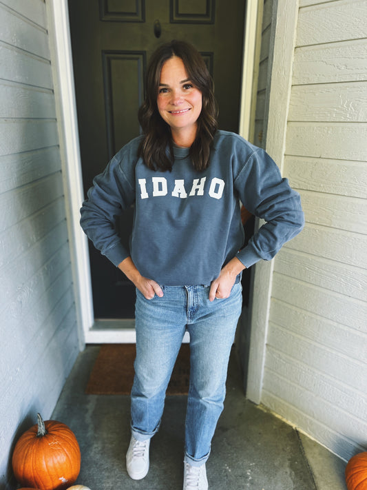 Person wearing a blue sweatshirt with 'Idaho' text, standing in front of a door with pumpkins on either side. The Idaho sweater is soft and cozy.