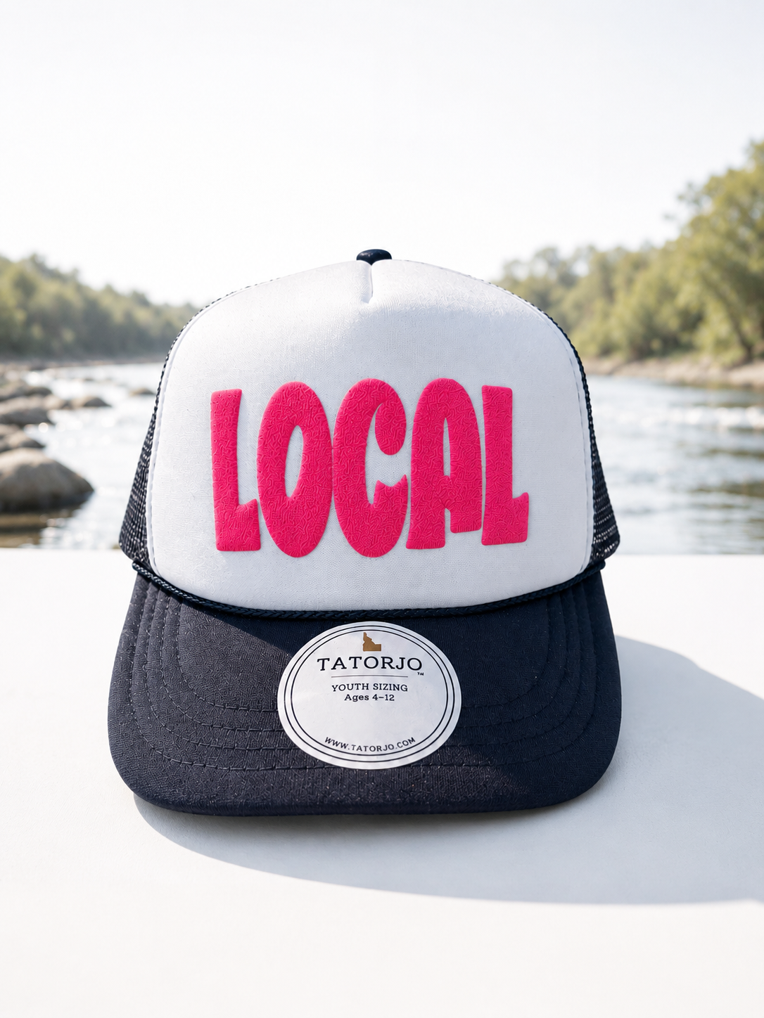 Trucker hat with 'LOCAL' in pink on a white background, featuring Tatorjo branding, with a natural landscape in the background.