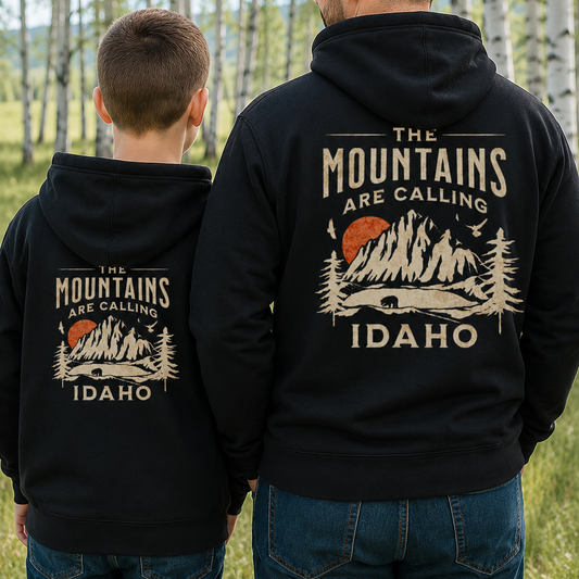 Two people wearing black hoodies with 'The Mountains Are Calling Idaho' design in a forest setting.