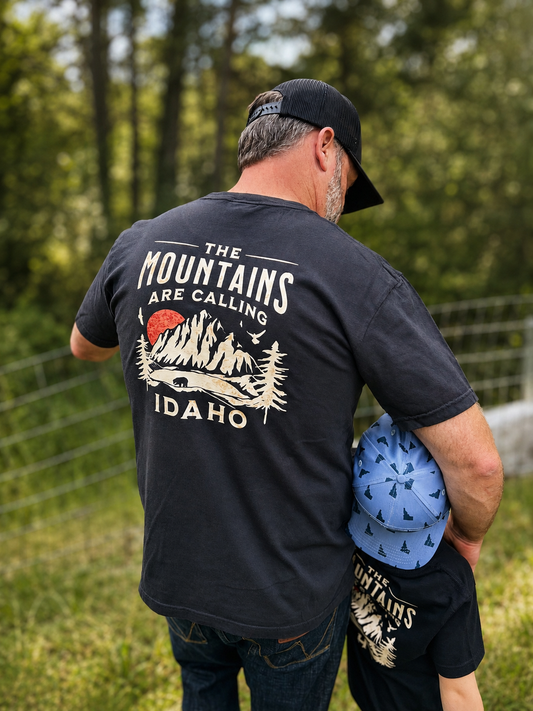 Man wearing a black t-shirt with 'The Mountains Are Calling Idaho' design, standing outdoors.