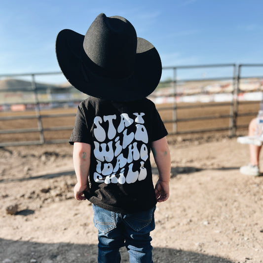 Child wearing a large cowboy hat and black t-shirt with text, the text says Stay Wild Idaho Child standing in an outdoor setting.