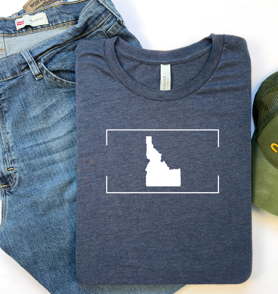 Navy blue Idaho long-sleeve shirt with a minimalist white Idaho outline design inside a rectangular frame, displayed as a flat lay with a hat next to it. This Idaho long sleeve t shirt is designed by TatorJo, an Idaho clothing company