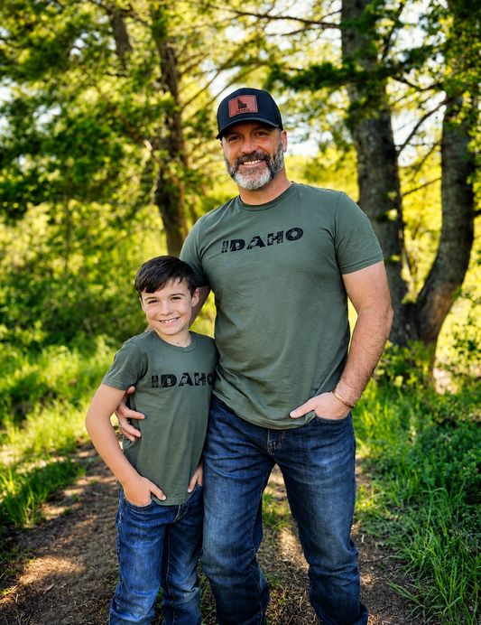 Man and young boy wearing Idaho t-shirts standing outdoors with trees in the background